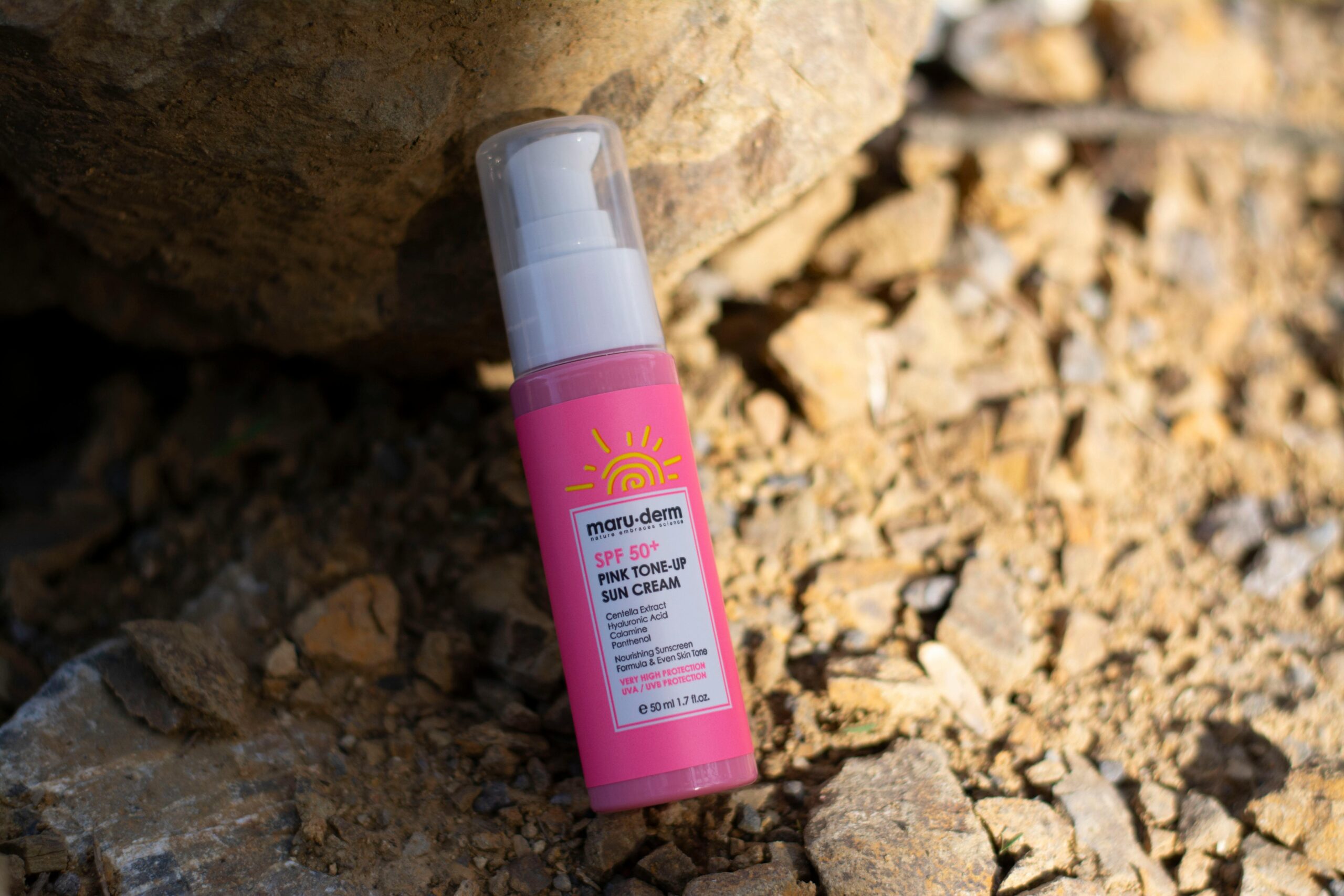 Close-up of SPF 50+ sun cream bottle with pink toned packaging on rocky surface.